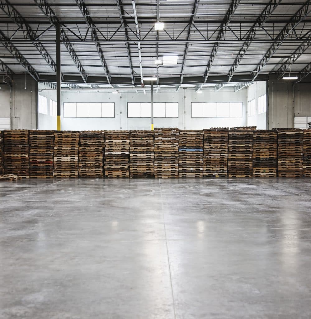 Interior of a warehouse with concrete floor, with a stack of pallets along the back wall.