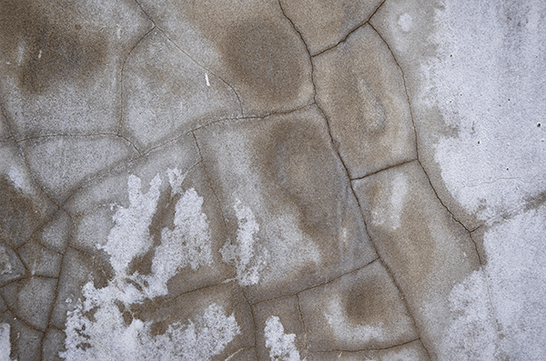 A top-down photo showing a cracked concrete floor covered in a light layer of snow.